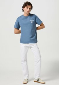 Camiseta de algodón azul con mangas cortas y cuello redondo, con un logo blanco de Wrangler. Combinada con pantalones blancos y zapatillas casuales.