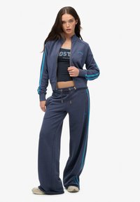Chándal para mujer de color azul marino con chaqueta con cremallera y pantalones holgados, con franjas laterales turquesas y un top corto ajustado debajo.