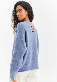 Niebieski puszysty sweter z okrągłym dekoltem, opuszczonymi ramionami i ściągaczami na mankietach. Na plecach znajduje się czerwona i czarna metka.