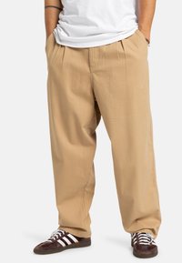 Beige broek met een ontspannen pasvorm, voorzien van voorplooien en zijzakken, gecombineerd met donkerbruine sneakers met witte strepen.