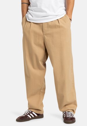 Beige broek met een ontspannen pasvorm, voorzien van voorplooien en zijzakken, gecombineerd met donkerbruine sneakers met witte strepen.