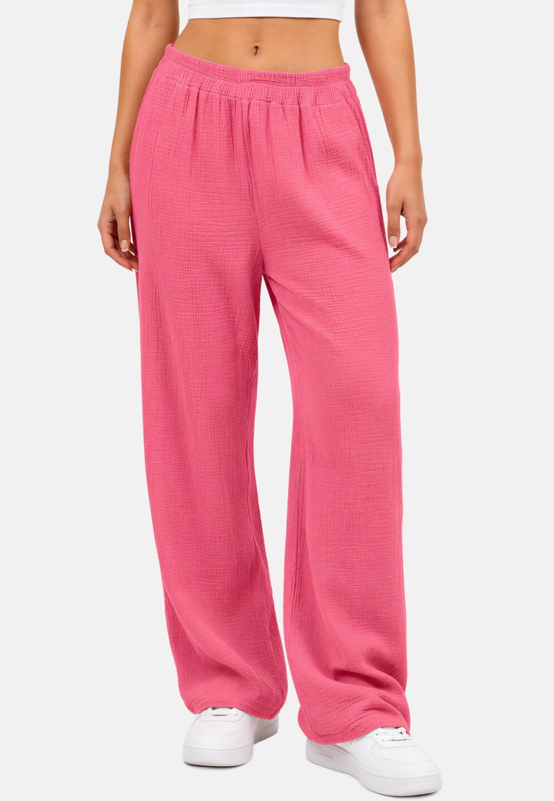 Vrouw die losse, gestructureerde roze broek draagt met elastische taille en witte sneakers, staand tegen een effen achtergrond.