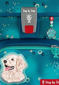 Teal backpack with a dog applique, featuring a silver logo clasp, colorful circular patterns, and a zippered pocket with paw print charm.