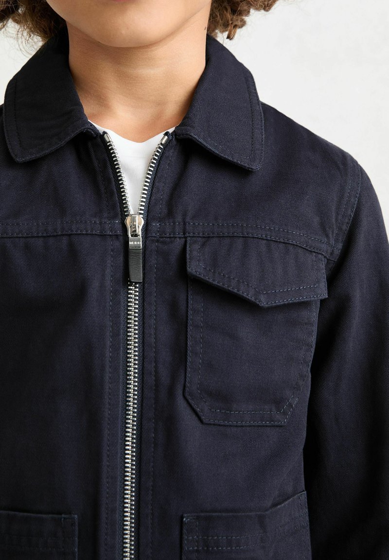 Veste en coton bleu marine avec une fermeture éclair à l'avant, un col pointu et deux poches poitrine. Présente des coutures visibles et un tissu texturé.