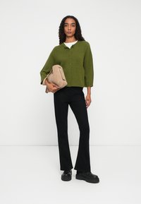 Kvinna som bär en grön knapp-up cardigan, svarta utsvängda byxor, svarta platåskor, och håller en beige handväska mot en enfärgad vit bakgrund.