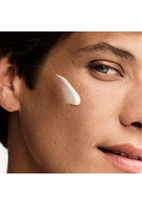 Vit kräm på ljusbrun hud, applicerad på kinden, med en närbild på ögat och läpparna. Slät textur, subtila detaljer synliga.