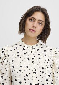 Blusa blanca con lunares negros, mangas cortas abullonadas, cierre frontal de botones y cuello alzado. Textura de tela suave.