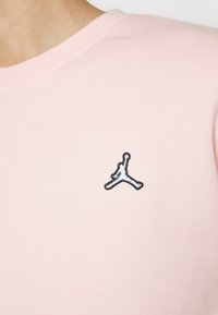 Camiseta de algodón rosa claro con cuello redondo, que presenta un logo Jumpman bordado en blanco y negro en el lado izquierdo del pecho.