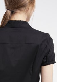 Camicia nera a maniche corte con polsini abbottonati, texture liscia e scollatura con colletto, che mostra la vista posteriore e dettagli di sartoria sottili.