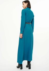 Abito teal a maniche lunghe con vita aderente, polsini bottonati e una cintura marrone, abbinato a stivali neri con tacco. Tessuto liscio, design lungo fino alla caviglia.