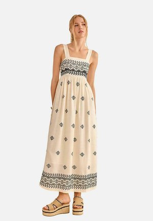 Mujer con un vestido maxi sin mangas color crema con patrones geométricos negros y sandalias de plataforma tejidas.