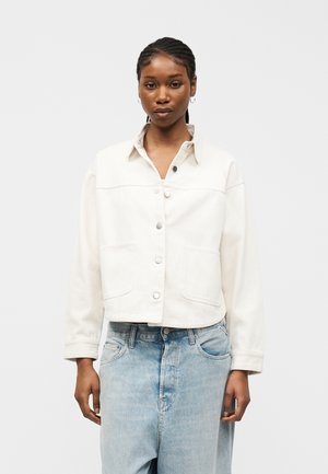 Γυναίκα με πλεξούδες στα μαλλιά, που φοράει λευκό σακάκι με κουμπιά και ανοιχτό μπλε τζιν, στέκεται μπροστά σε ένα απλό φωτεινό φόντο.