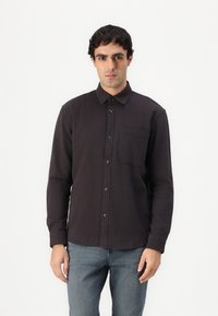 Camicia a maniche lunghe di colore scuro con un'unica tasca sul petto, bottoni neri e una texture uniforme. Indossata con jeans grigi a contrasto.