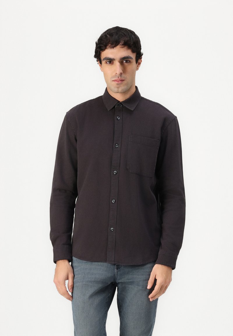 Camicia a maniche lunghe di colore scuro con un'unica tasca sul petto, bottoni neri e una texture uniforme. Indossata con jeans grigi a contrasto.