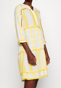 Vestido blanco con rayas amarillas y patrones de hojas. Presenta una tapeta de botones, mangas de tres cuartos y un dobladillo con volantes. Hecho de una tela ligera.