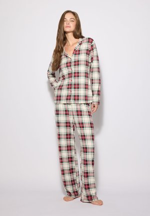 Donna in piedi a piedi nudi che indossa una camicetta a quadri rossa, nera e bianca con bottoni e maniche lunghe e pantaloni abbinati su sfondo uniforme.