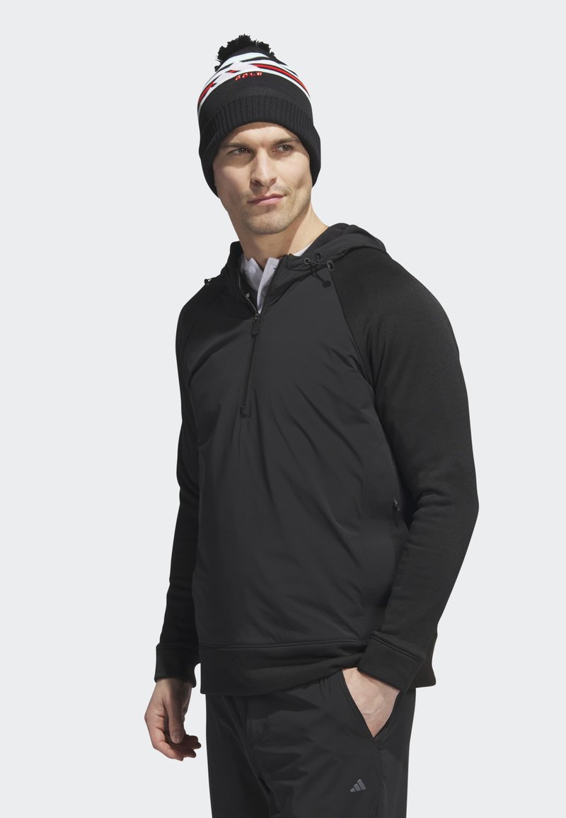 Schwarzer Pullover-Hoodie mit Reißverschluss am Kragen, langen Ärmeln und einer Kapuze mit Kordelzug. Getragen mit einer schwarzen Strickmütze, die rote und weiße Akzente hat.