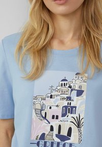 T-shirt bleu clair avec un imprimé graphique d'un paysage grec, comprenant des bâtiments blancs et des dômes bleus. Manches courtes, texture douce.