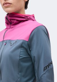 Gros plan d'une personne portant une veste rose et grise avec un logo sur la poitrine et « DYN » imprimé sur la manche.