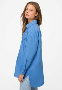 Light blue oversized shirt with a collar, single chest pocket, and side slits. Features a design element on the back in contrasting color.