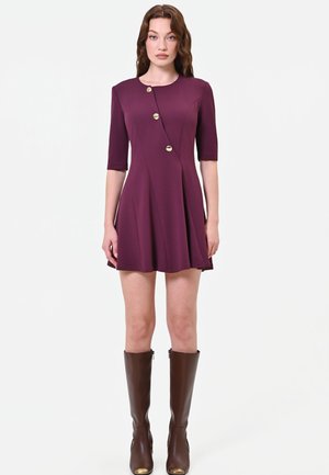 Femme portant une robe prune longueur genou avec trois boutons dorés et des manches arrivant aux coudes, associée à des bottes marron montant aux genoux.