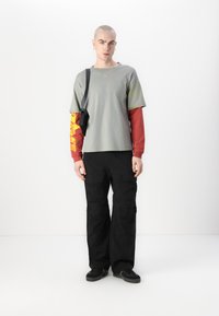 T-shirt gris superposé sur une chemise à manches longues rouge avec des graphiques jaunes, associé à un pantalon cargo noir et des baskets noires.