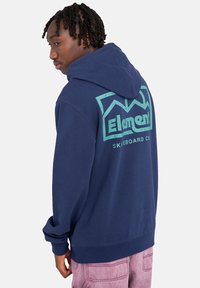 Sudadera con capucha azul marino con un gran logotipo gráfico verde en la parte posterior, que presenta un ajuste holgado y puños y dobladillo acanalados.