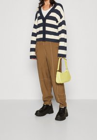 Randig marinblå och gräddvit kofta med knappar, tillsammans med bruna utility-byxor med vit sömnad och svarta plattformsskor. Gul handväska.