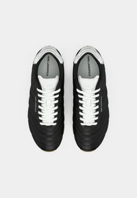 Ténis pretos em couro texturizado, com atacadores brancos e marca Karl Lagerfeld na palmilha cinzenta e na língua. Apresentam um design com biqueira arredondada.