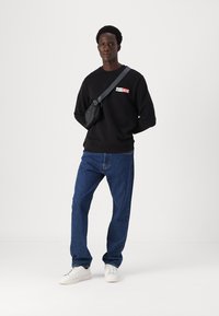 Svart hettegenser med rød og hvit merkevare, blå denimjeans og hvite joggesko. En liten svart crossover-veske bæres over kroppen.