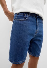 Pantalones cortos de mezclilla azul con un ajuste regular, que cuentan con cremallera, cinco bolsillos y costuras visibles. El material tiene un acabado suave y texturizado.
