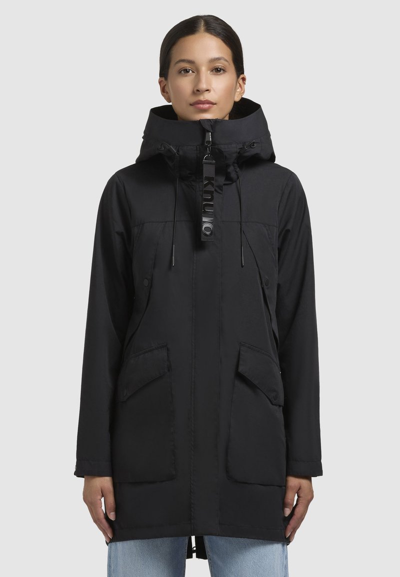 Parka noire avec un col haut, une capuche réglable et deux poches avant. Fabriquée en tissu léger avec un léger brillant.