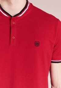 Röd polotröja i texturerat tyg, med en svart sköld, två knappar och en ribbad krage med vita och svarta ränder.