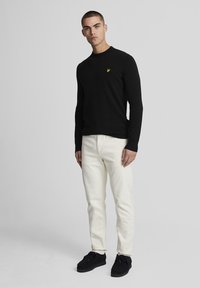 Fekete hosszú ujjú pulóver sárga logóval, fehér farmerrel és fekete, bőrszínű velúr cipővel párosítva. Texturált anyag és testhezálló szabás.