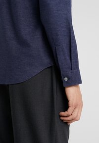 Rechterarm en hand van een persoon die een marineblauwe lange mouw shirt met een knoopmanchet en donkergrijze broek draagt tegen een effen achtergrond.