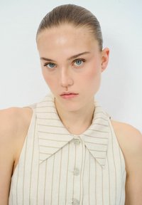 Lichtbeige mouwloze shirt met een grote kraag, verticale strepen en knoopdetails. Gladde textuur en gestructureerd ontwerp.