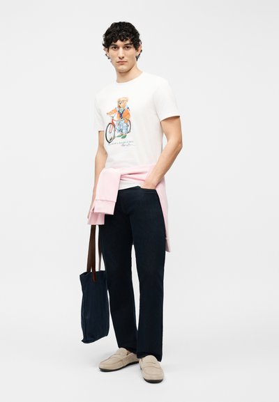 Άνδρας που φοράει λευκό μπλουζάκι με στάμπα αρκούδας σε ποδήλατο, μαύρο παντελόνι, μπεζ μοκασίνια, ροζ πουλόβερ δεμένο στη μέση, κρατώντας τσάντα ώμου σε ναυτικό μπλε χρώμα.