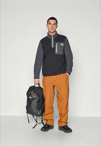 Sort og grå fleecepullover med kvart lynlås og brystlomme, parret med orange cargo bukser og en sort rygsæk. Sorte sko.