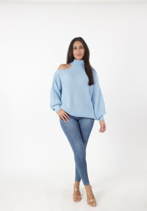 Lichtblauwe gebreide trui met een off-shoulder ontwerp, gecombineerd met skinny blauwe jeans en transparante haksandalen.