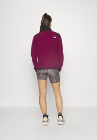 Fleecejacka i borgogne med svarta accenter, kombinerad med mönstrade shorts och träningsskor. Strukturerat material, figurnära design.