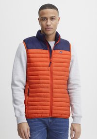 Polstret vest i lys orange med en mørkeblå krave og lynlås, med lodrette quiltede sektioner og to sidelommer.