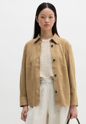 Woman with long black hair wearing a beige suede button-up jacket, cream knit sweater, white pants, and holding a brown handbag.