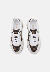 Zapatilla blanca con paneles de textil negro que presentan un patrón dorado, punta redonda y cordones blancos. Logotipo impreso en la plantilla. Suela de goma.