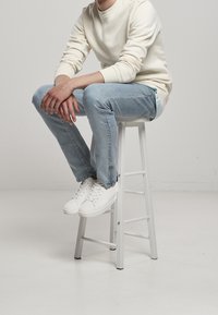 Taburete de madera blanca con una figura sentada que lleva una sudadera crema, jeans azul claro y zapatillas blancas. Texturas suaves; diseño casual.