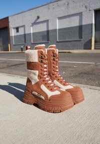 Bottes hautes en peau lainée marron et blanche à lacets avec des semelles épaisses, posées sur un trottoir en ville près d'une rue et de bâtiments.
