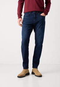 Dark blue denim jeans with a straight leg fit, stitched detailing, and pockets, paired with tan suede ankle boots.