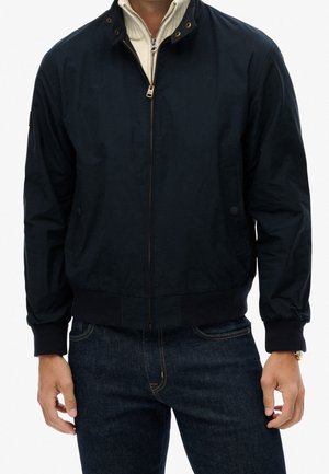 Homme portant une veste zippée bleu marine foncé par-dessus un pull zippé crème clair, associé à un jean bleu foncé.
