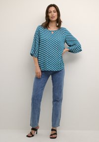 Bluză teal cu un model alb în zig-zag, detaliu chei, și mâneci trei sferturi, purtată cu blugi drepți albaștri și sandale cu toc negre.