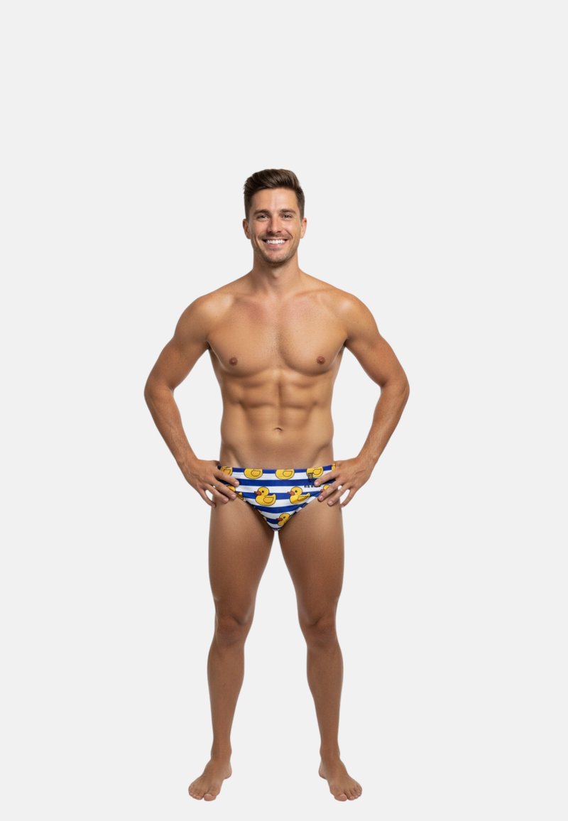 Uśmiechnięty muskularny mężczyzna stojący boso, ubrany w niebiesko-białe prążkowane kąpielówki z nadrukiem żółtych gumowych kaczek na białym tle.