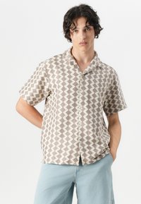 Short-sleeved shirt in beige fabric featuring a geometric brown pattern, button-front closure, and a collar. Paired with light blue shorts.